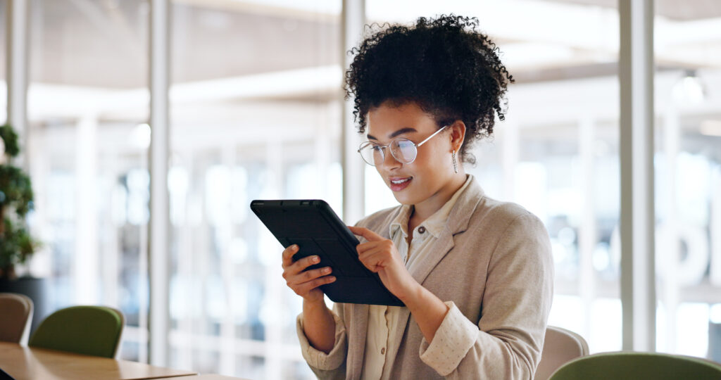 Frau mit lockigem Haar und Brille studiert konzentriert an einem Tablet in einem modernen Büro, das einen Verhaltenskodex zeigt, im Hintergrund sind unscharfe Büromöbel und Pflanzen zu sehen.